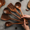 Main tenant un lot de spatules et cuillères en bois sombre, posées sur un fond de table rustique.