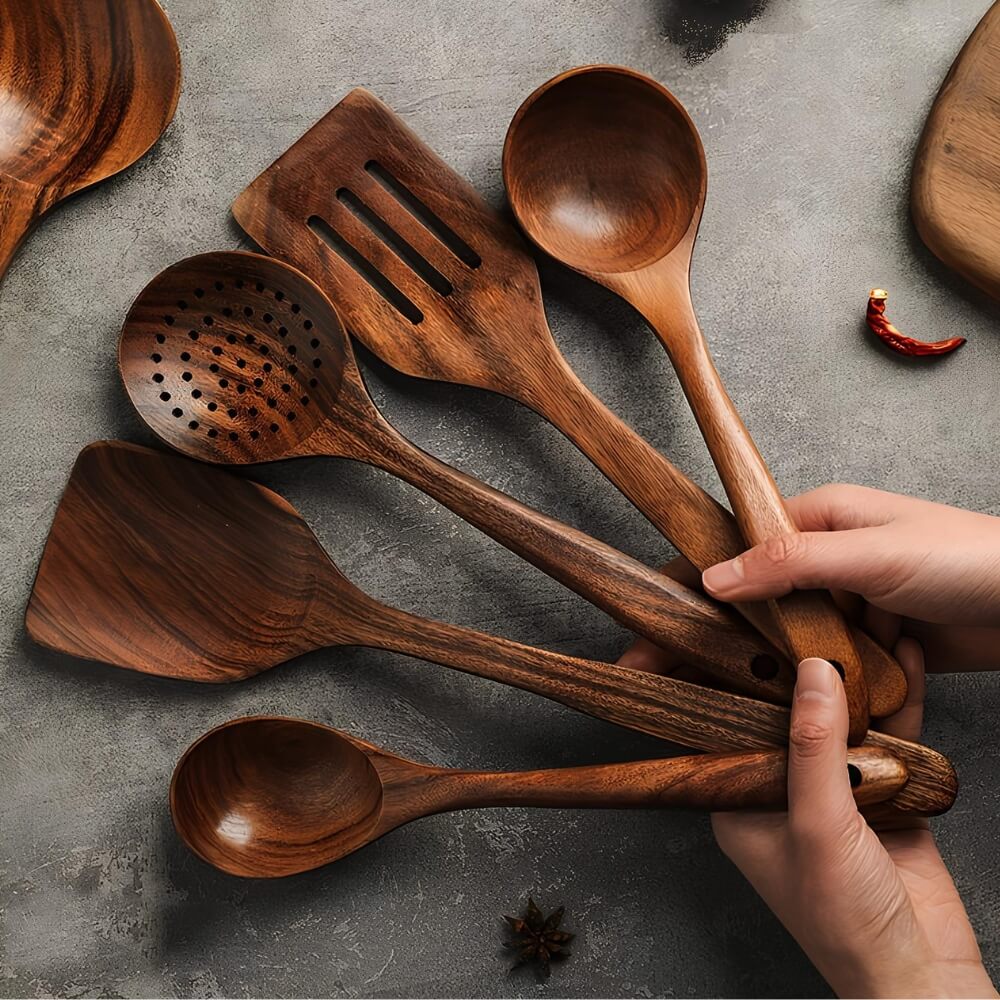 Main tenant un lot de spatules et cuillères en bois sombre, posées sur un fond de table rustique.