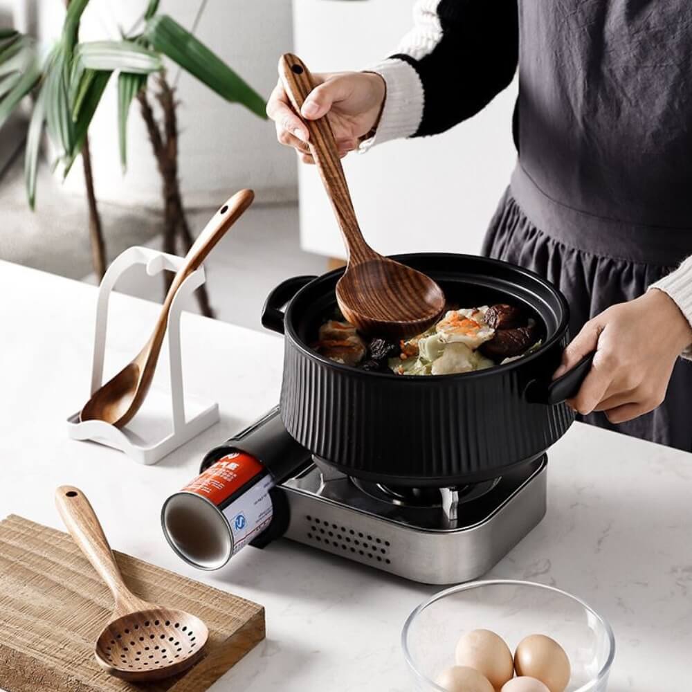 Louche en bois servant un potage aux légumes dans une marmite noire sur réchaud à gaz portatif.