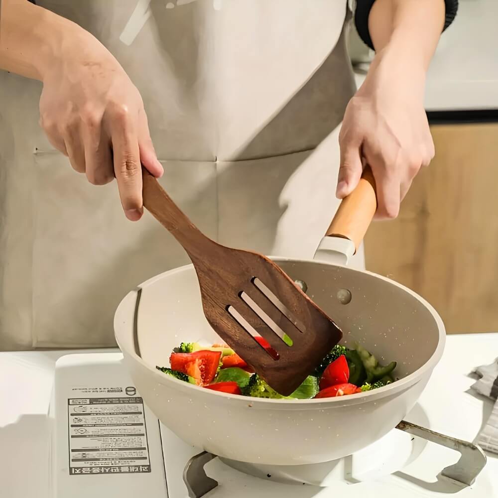 Personne cuisinant des légumes frais dans une poêle à l’aide d’une spatule ajourée en bois.