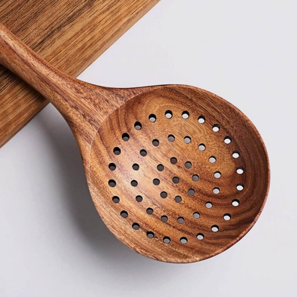 Cuillère écumoire en bois naturel avec trous réguliers, posée sur une planche en bois clair.