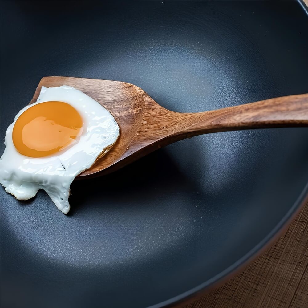 Spatule en bois soulevant un œuf au plat dans une poêle noire antiadhésive.
